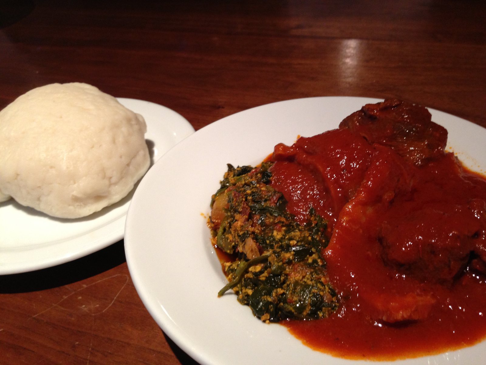 A mound of pounded yam (iyan) on a white plate, served beside a plate of red stew and egusi soup with greens