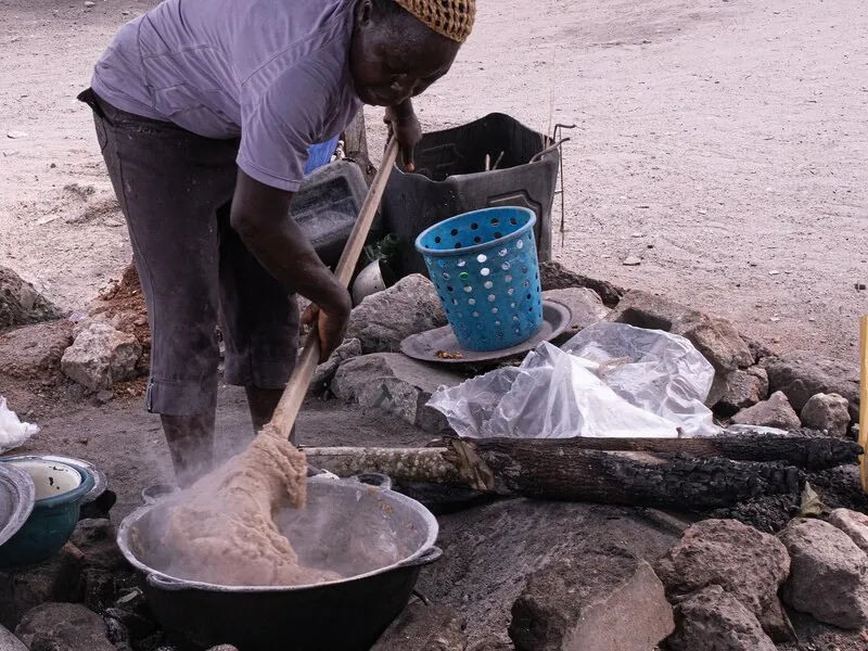 A woman turning a steaming yam-flour dough in a pot over a fire with a long wooden stick